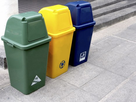 In the foreground, there are three upright wheeled rubbish bins positioned on a paved outdoor area adjacent to a building or street. The first bin on the left is green with a matte finish and a rounded lid, featuring a small white triangular recycling symbol on its side. The middle bin is yellow with a slightly textured surface, also with a rounded lid, and displays a black recycling symbol on its front. The third bin on the right is dark blue, with a smooth, slightly glossy finish, and has a rectangular white label or logo on its front side. The bins are arranged in a line, with the yellow bin slightly behind the green one and the blue bin placed at an angle, touching the yellow bin. The pavement is composed of grey concrete slabs, and there is a shadow cast by the bins indicating daytime lighting. The setting appears to be outside, possibly near an office or commercial premises in Docklands, fitting within the scope of rubbish collection and waste management services offered by Office Clearance Docklands.