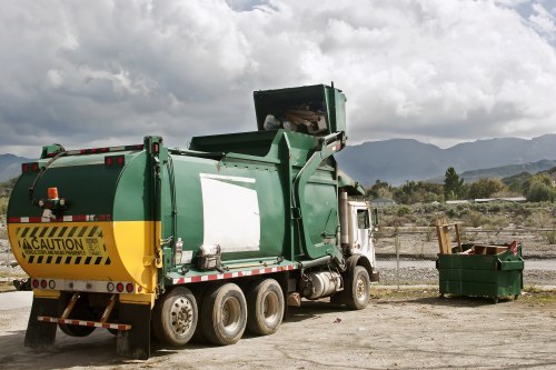 A large green refuse collection truck with a white cabin is parked on a gravel surface outdoors. The truck has a yellow and black hazard caution sticker on the rear and is equipped with a waste compaction mechanism at the back, which appears to be slightly raised. To the right of the truck, there is a green wheelie bin filled with various waste materials, situated on the ground. In the background, a chain-link fence runs along the perimeter, and beyond the fence, there are hilly landscapes with sparse vegetation and distant mountains under a partly cloudy sky. The scene is consistent with a rubbish collection or waste disposal operation, likely within a commercial or industrial yard, typical of areas near Docklands or similar urban outskirts, as handled by a company like Office Clearance Docklands specializing in rubbish removal services.