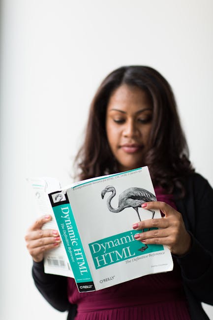 A woman with dark, wavy hair wearing a black blazer and a maroon top is holding open a book titled 'Dynamic HTML' by Oreilly, which features an illustration of a flamingo on the cover. She is examinin