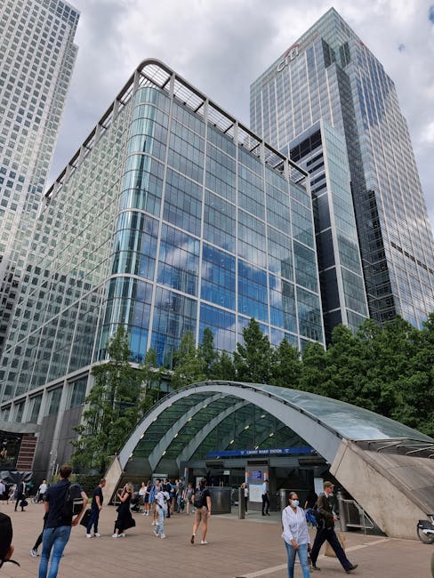 A night-time view of the Canary Wharf financial district skyline, featuring tall, modern office buildings with illuminated windows. The buildings have a variety of architectural styles and are reflect
