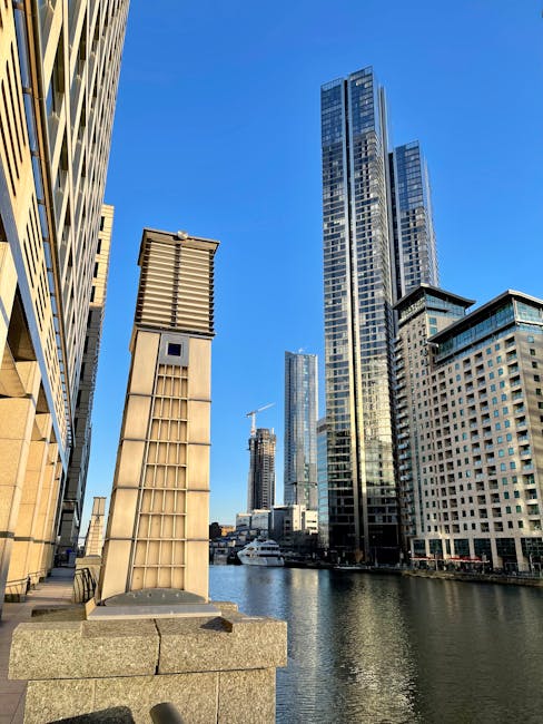 A panoramic cityscape of modern high-rise buildings and office towers in Docklands, with some structures featuring glass facades reflecting the warm light of the setting sun. Several cranes are visibl