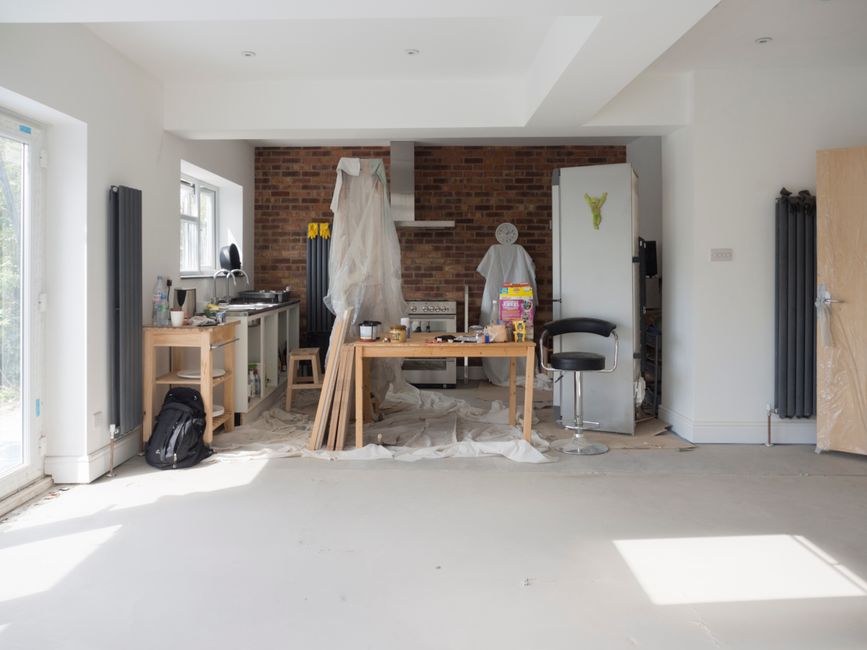 The image depicts a partially assembled white-painted wooden framed window or door with multiple glass panes, leaning against a plain light grey internal wall. Two smaller similar frames are positione