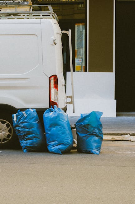 The image shows the rear side of a white commercial van parked beside a building with large glass windows and a dark frame. Three blue heavy-duty refuse bags, made of thick plastic with a slightly cri