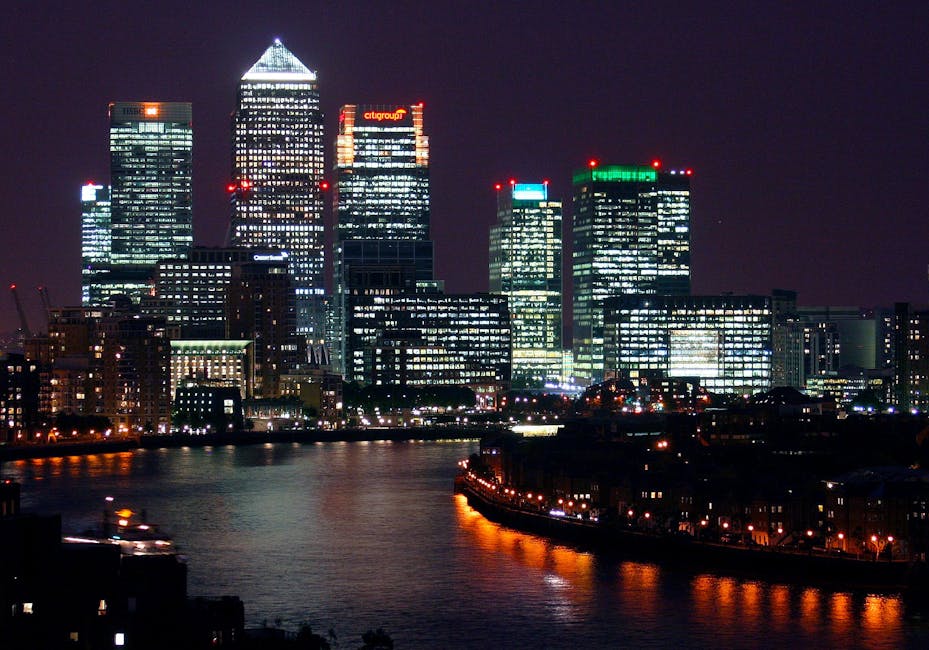 A wide view of a modern urban waterway flanked by high-rise buildings with varied architectural styles and materials, including glass, concrete, and brick facades. The foreground shows calm, reflectiv